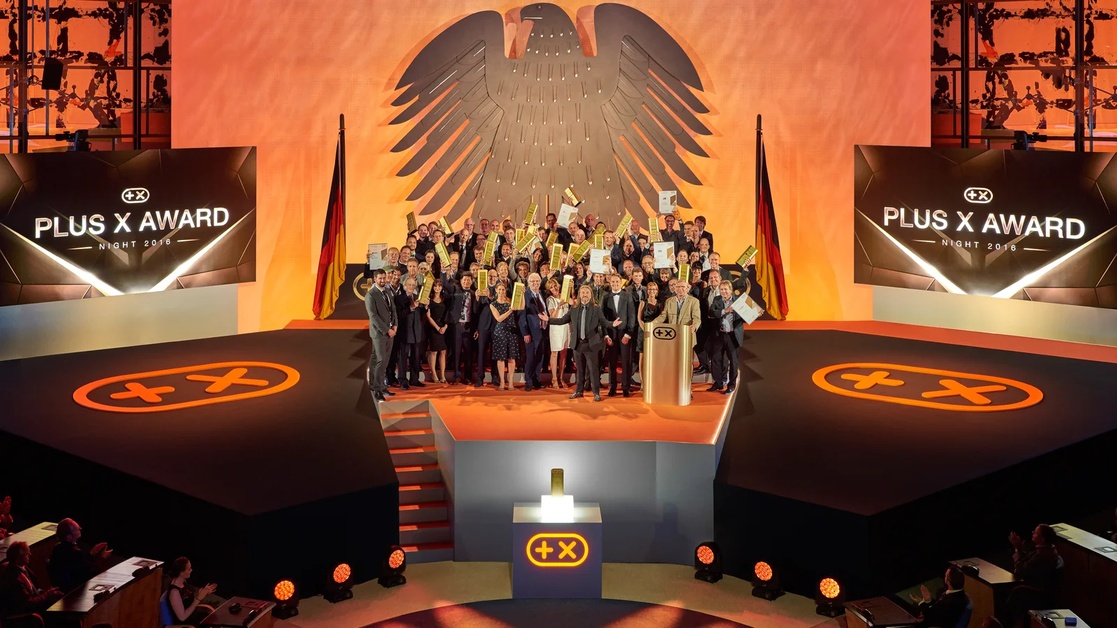 Bühne der PLUS X AWARD Night: rund achtzig Ausgezeichnete stehen mit ihren Trophäen unter dem Bundesadler, links und rechts Anzeigetafeln mit dem PLUS-X-AWARD-Zeichen