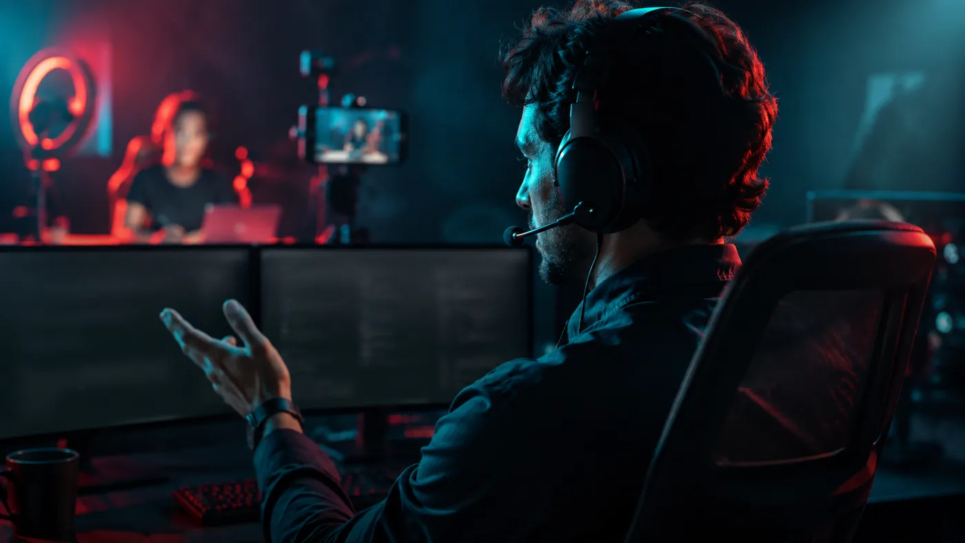 Mann mit Headset gestikuliert vor zwei Bildschirmen, im Hintergrund ein Streaming-Aufbau