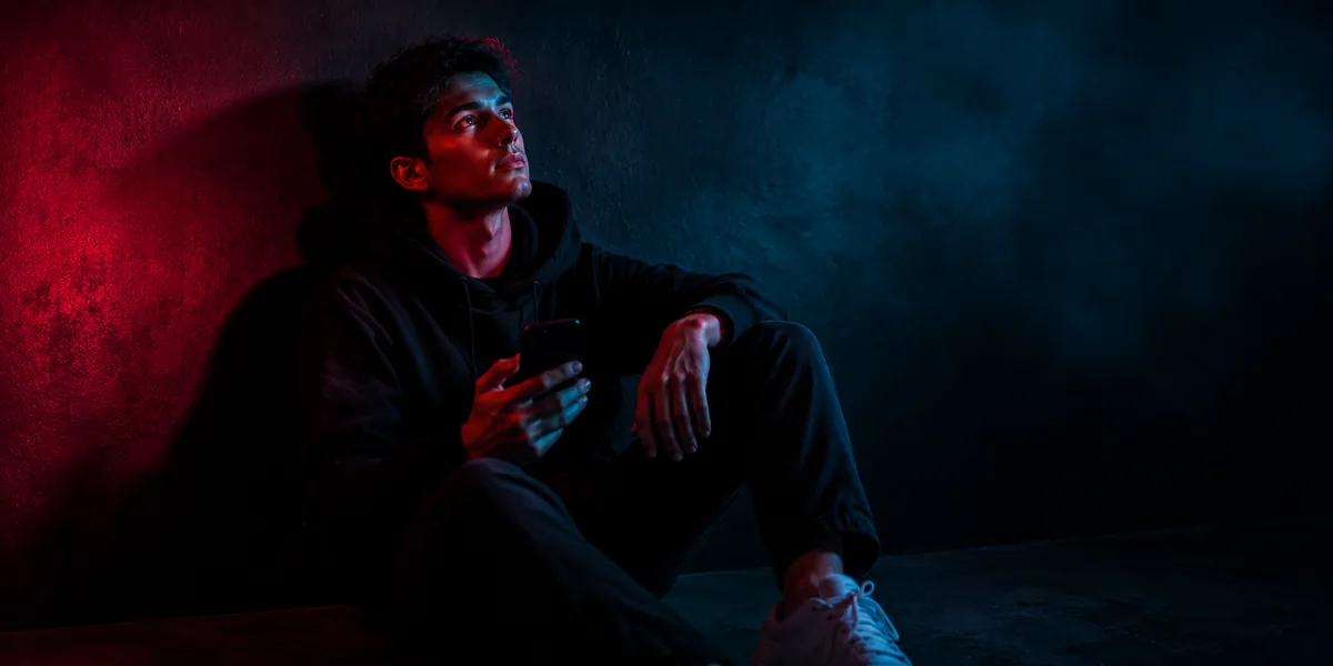 Junger Creator sitzt mit dem Handy in der Hand an einer Wand und blickt nach oben